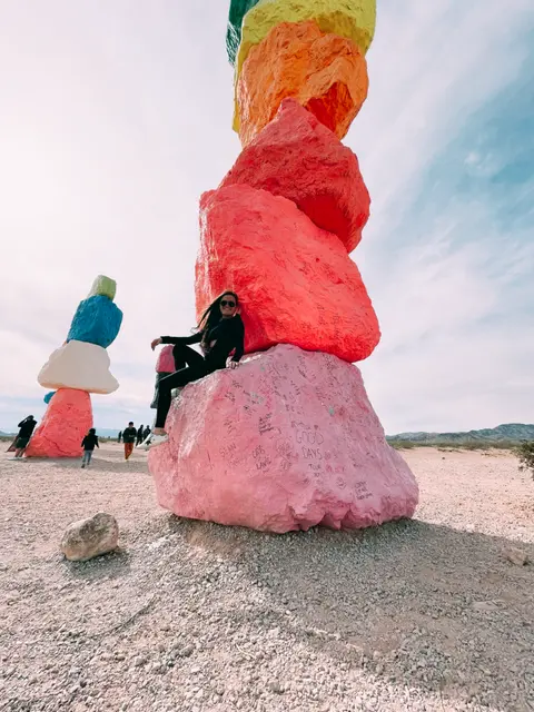 A woman poses sitting on a large, brightly painted boulder with other colorful rock formations in the desert.