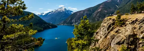 A scenic view of a blue mountain lake surrounded by forested mountains and a rocky cliff.