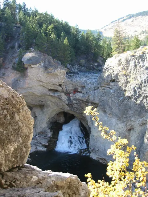 A waterfall flows through a natural rock arch surrounded by trees and cliffs.