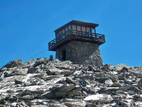 A stone and wood lookout tower stands on a rocky mountain peak under a clear blue sky.