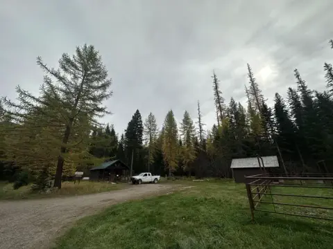 A white pickup truck parked in a clearing near small buildings and tall trees under an overcast sky.