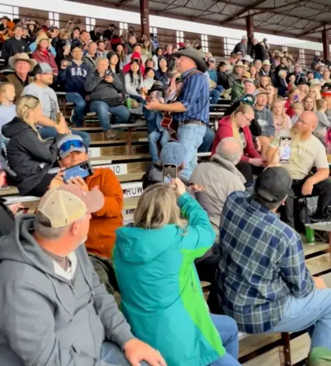 A man playing guitar in front of a large crowd in bleachers.