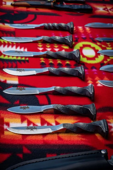 Several handmade knives with handles made from railroad spikes are laid out on a patterned red blanket.