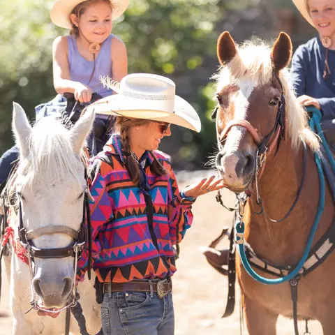A woman stands between two horses being ridden by children outdoors.