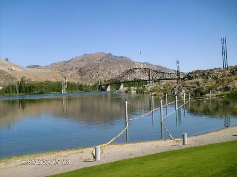 A river flows past a bridge beneath arid hills under a blue sky.