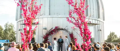 A couple participates in an outdoor wedding ceremony framed by vibrant floral arrangements.