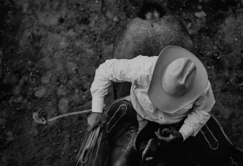 A high-angle black and white photograph of a person in a cowboy hat riding a horse and holding a rope.