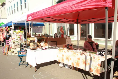 A vendor sits inside a red canopied booth at an outdoor market.