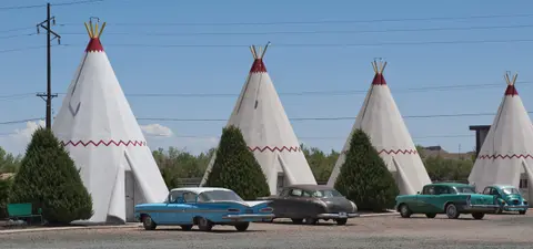 A row of white teepee-shaped motel units with vintage cars parked in front.