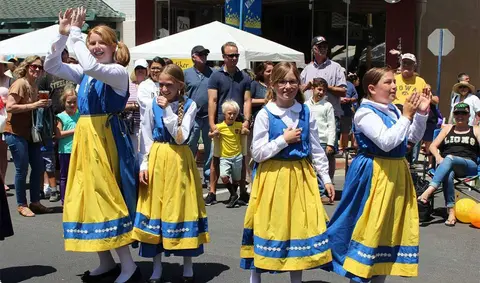 Four girls in traditional blue and yellow folk costumes perform outdoors in front of a crowd.