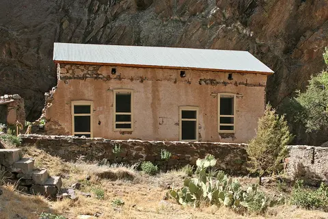An old adobe building with a corrugated metal roof stands against a rocky cliff face.