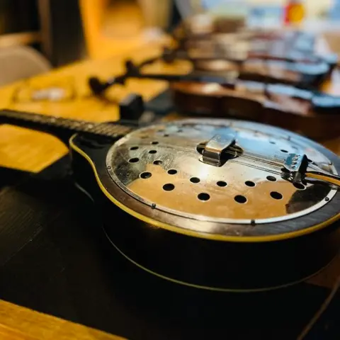 A resonator instrument rests on a wooden table with several violins in the background.