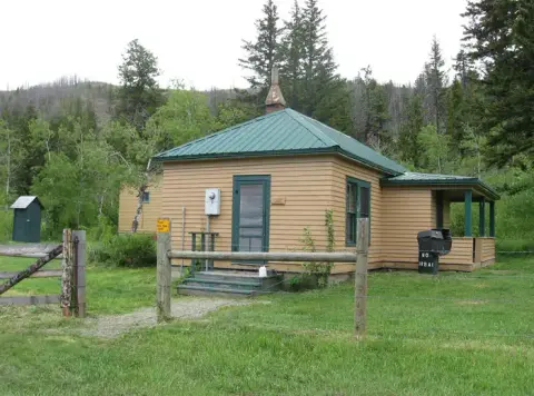 Exterior view of a small historic cabin with a green metal roof surrounded by trees and grassy land.