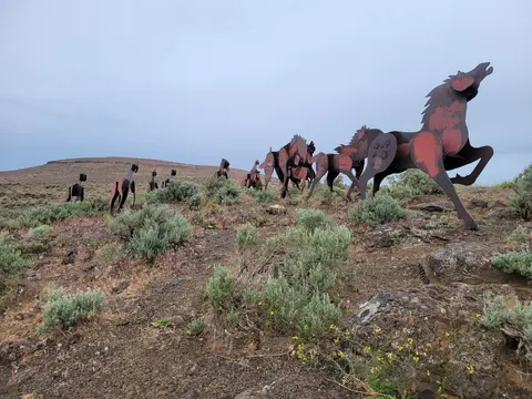 A line of metal horse sculptures stands on a barren hillside under an overcast sky.