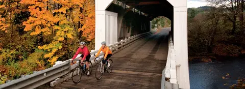 Two cyclists ride across a covered wooden bridge during autumn.