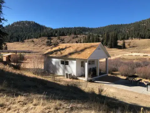 A small white building stands in a valley with mountains and trees in the background.