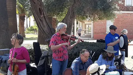 A small group of musicians play acoustic instruments outdoors near trees and a building.