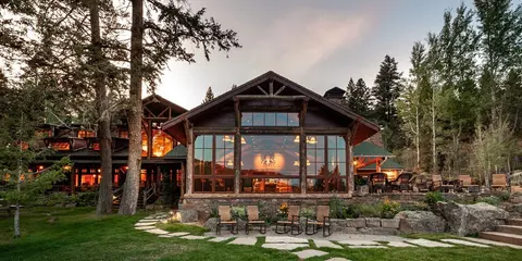 A large rustic cabin with glass walls and stone details is surrounded by trees.
