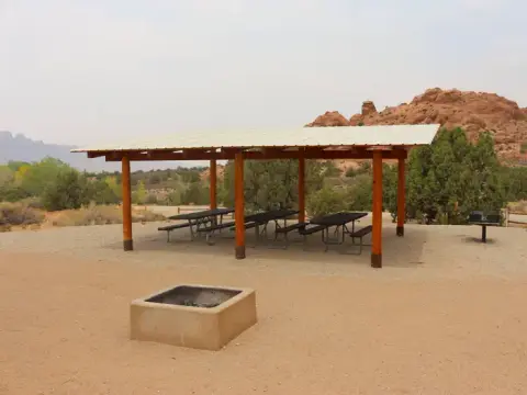 A covered picnic shelter with tables and a fire ring in a desert setting.
