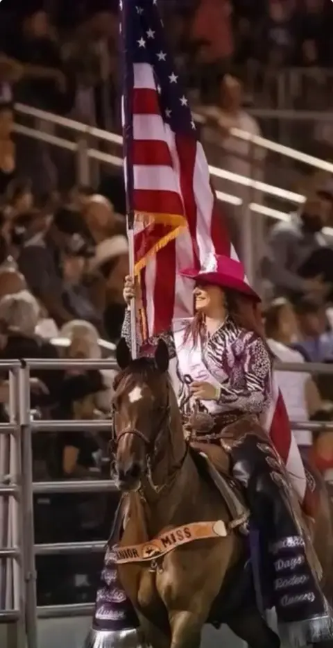 A cowgirl on a horse carries an American flag in an arena.