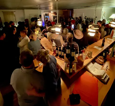 A crowded indoor bar with people gathered around wooden counters and bottle displays.
