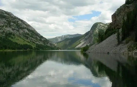 A scenic view of a calm lake surrounded by mountains and forests under a cloudy sky.