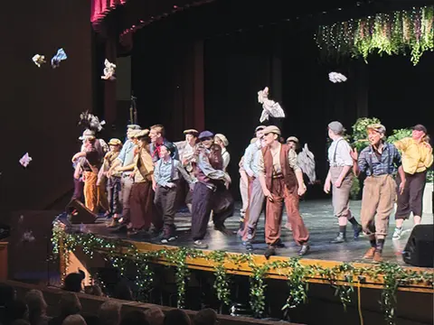 A group of performers act on a theater stage as papers fly through the air.