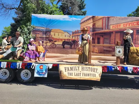 A parade float features people dressed in historical period costumes against a western town backdrop.