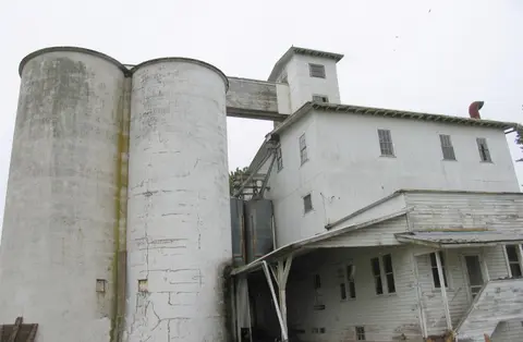 An exterior view of an old white industrial mill and silos under an overcast sky.