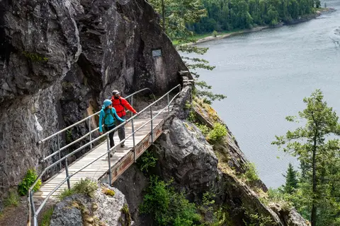 Two hikers walk along a wooden path built into a cliff side next to a river.