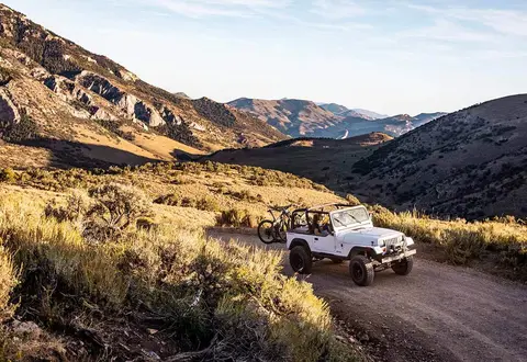 A white off-road vehicle carrying bicycles drives along a dirt road in a mountainous landscape.