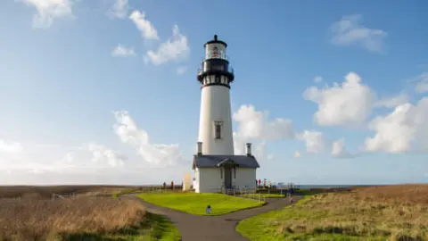 A tall white lighthouse stands in a grassy landscape under a blue sky with clouds.
