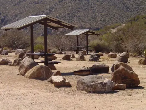 A desert campground features shaded picnic tables and fire rings surrounded by rocks.