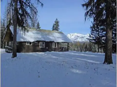 A log cabin covered in snow sits among trees with snowy mountains in the distance.