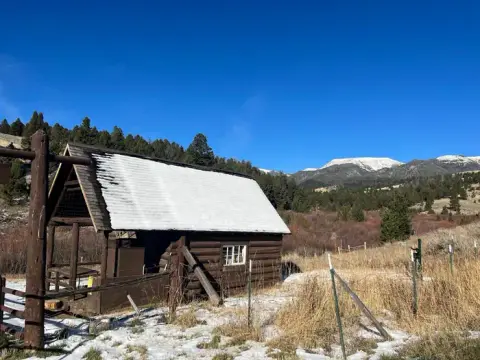 A small log cabin with snow on the roof sits in a grassy field with mountains in the background.