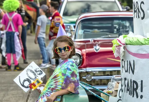 A young girl in a tie-dye shirt and peace-sign glasses holds a 60s sign at an outdoor event.