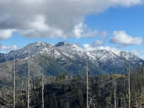 Snow-capped mountains rise above a forested landscape under a cloudy sky.