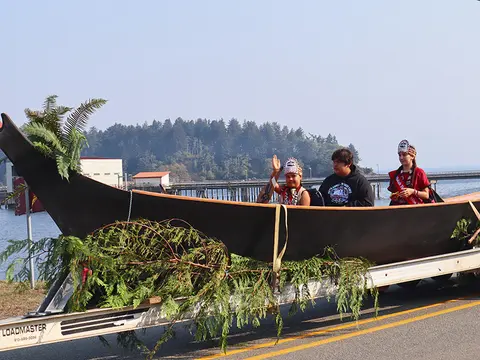 People sit inside a canoe decorated with greens on a trailer near the water.