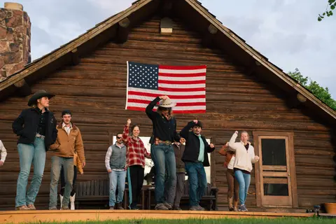 A group of people dancing outside a log cabin with an American flag hanging on the wall.