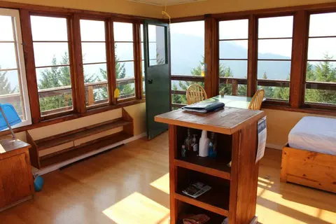 An interior view of a wooden fire lookout cabin with panoramic windows and a bed.