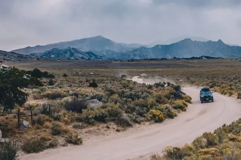 A vehicle drives along a dirt road through a desert landscape with mountains in the background.
