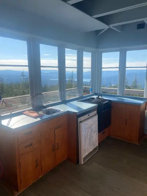 An interior view of a room with large windows overlooking a mountainous landscape, featuring a kitchenette with wooden cabinets and a stove.