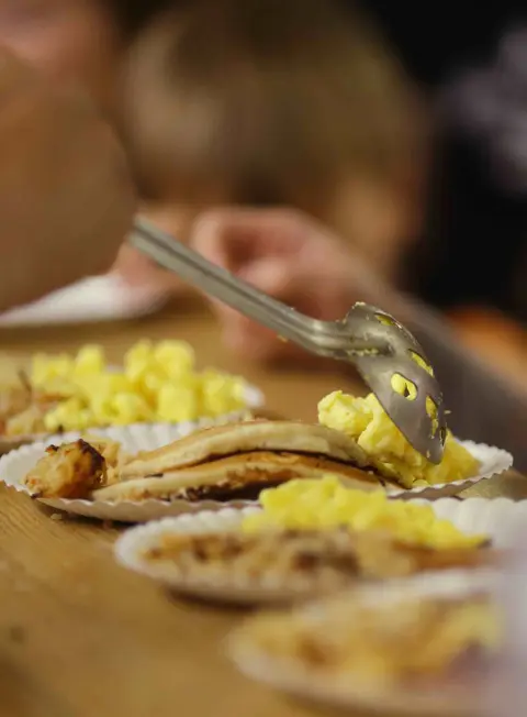 A person uses tongs to serve scrambled eggs onto paper plates containing pancakes.