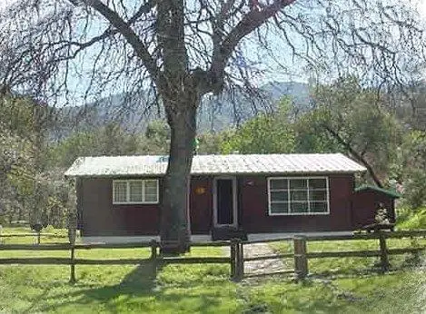 A one-story cabin with a white roof behind a wooden fence and large tree.