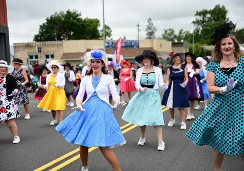 A group of women in vintage dresses and hats dance together in a street.
