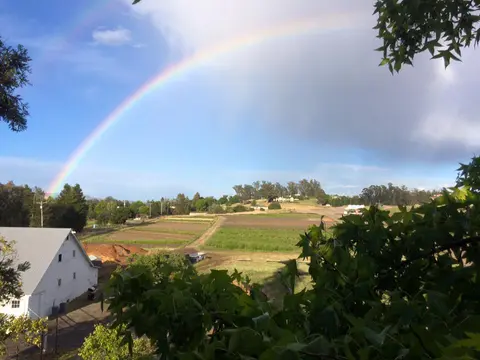 A large rainbow arches across the sky over a rural landscape with fields and a white building.