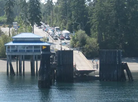An elevated view of a ferry terminal with a line of waiting cars on the road and a wooden pier structure leading to the water.