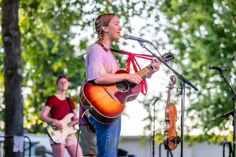 A young woman plays an acoustic guitar and sings into a microphone outdoors.