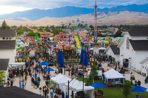 An elevated view of a crowded county fair with rides, booths, and mountains in the background.
