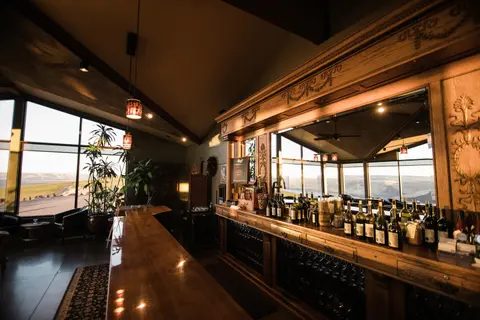 An interior view of a stylish wooden bar with bottles lined up and large windows showing a view.